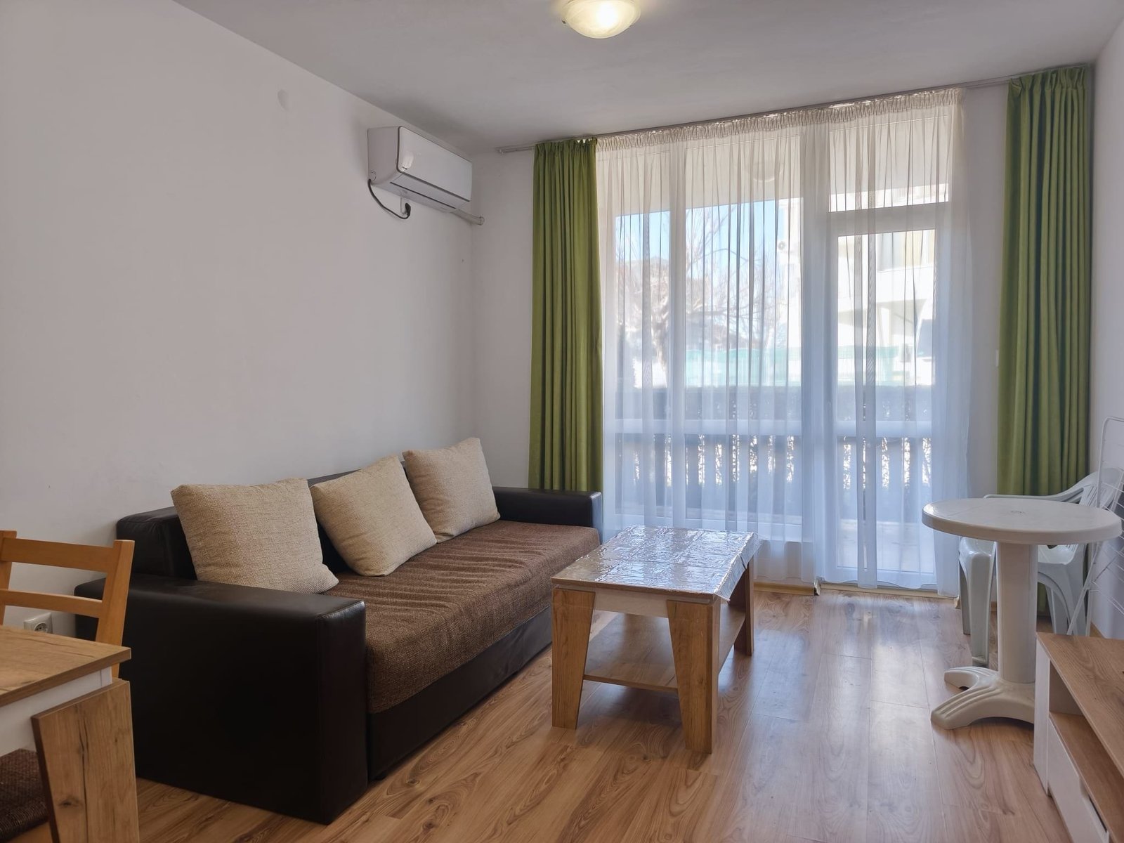 Современная квартира с уютной гостиной и большим окном Современная гостиная в Болгарии с диваном, столом и большими окнами, создающими светлую и уютную атмосферу.