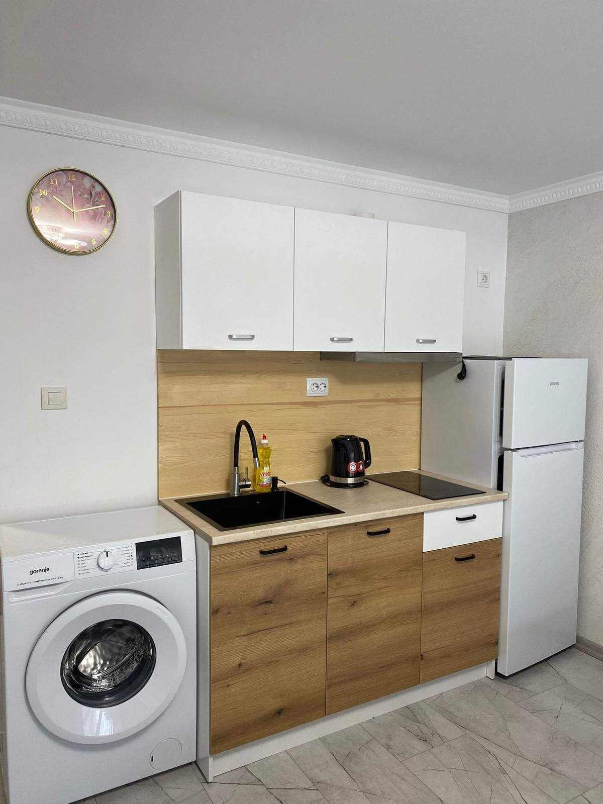 Современная кухня с гладкими белыми шкафами, стиральной машиной, холодильником и черной раковиной в интерьере Болгарской недвижимости.