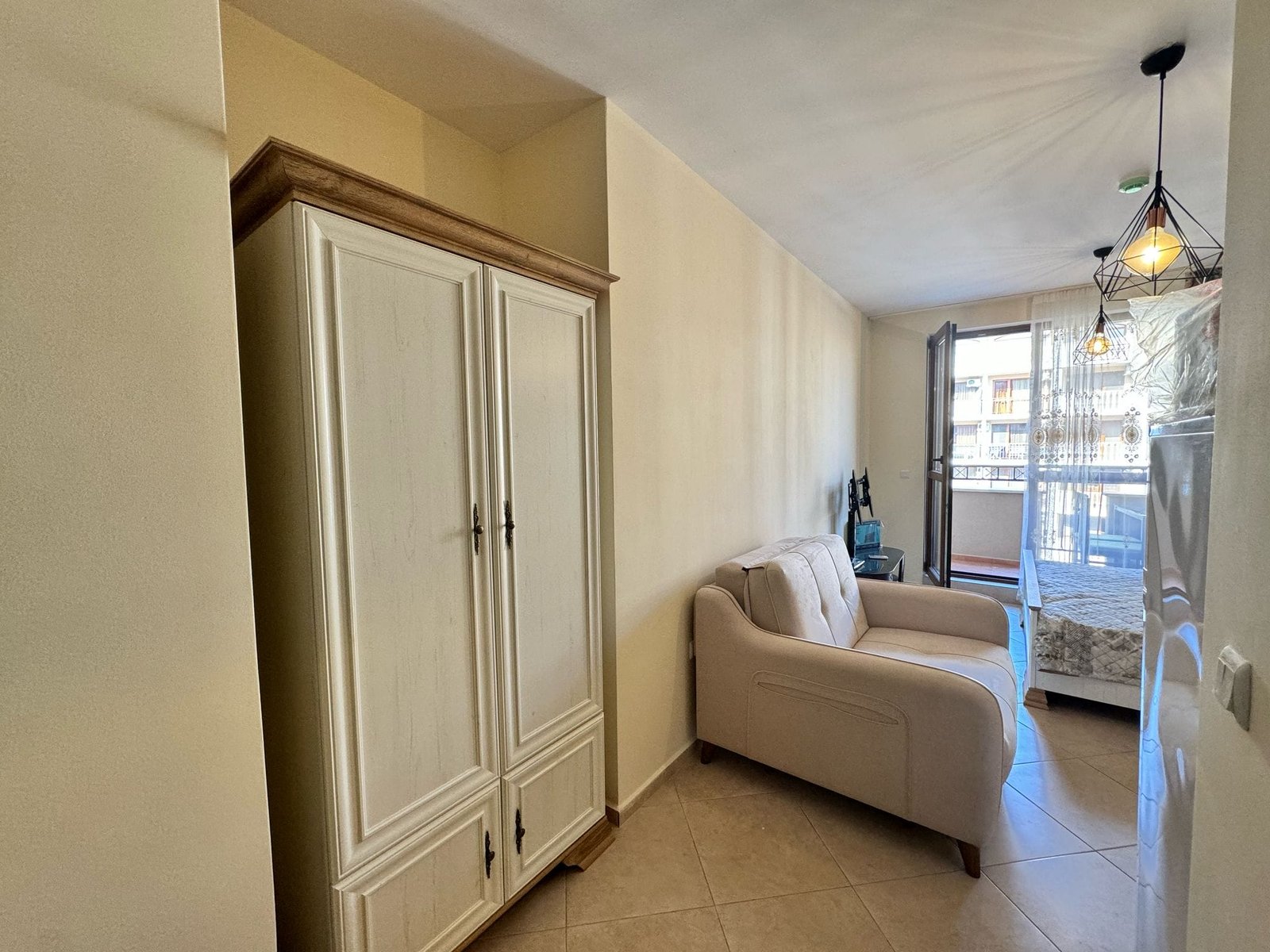 Светлая комната с диваном и шкафом, выход на балкон, идеальное предложение для покупки недвижимости в Болгарии.