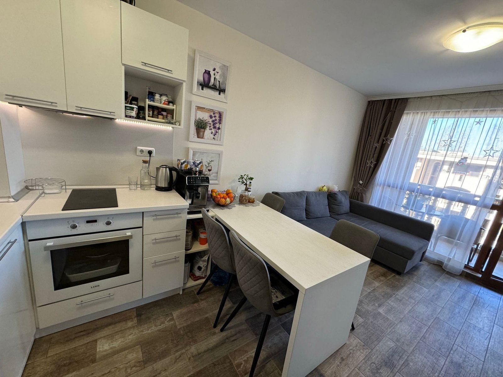 Современная кухня-гостиная в светлых тонах с диваном и балконом Современная светлая кухня с обеденным столом, уютный уголок для отдыха, окна с видом на улицу, элементы дизайна в белых тонах.