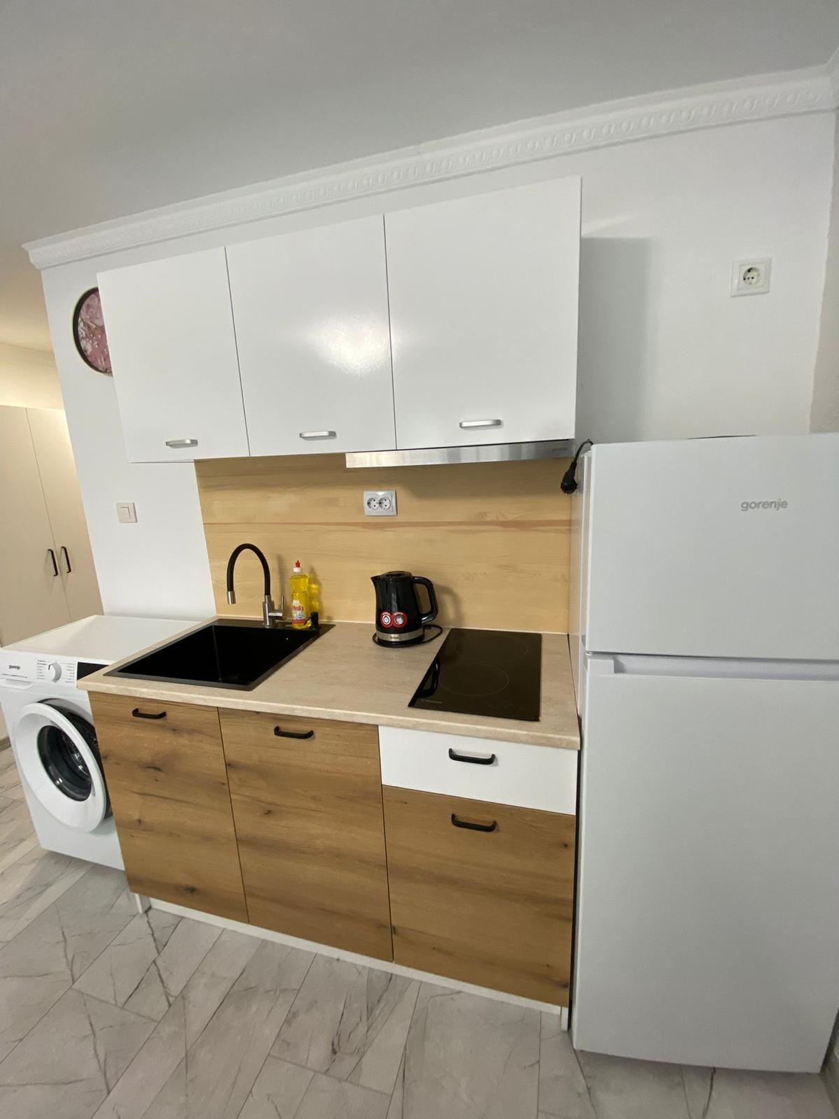 Современная кухня с белыми и деревянными фасадами, встроенной техникой и стиральной машиной, идеально для покупки недвижимости в Болгарии.