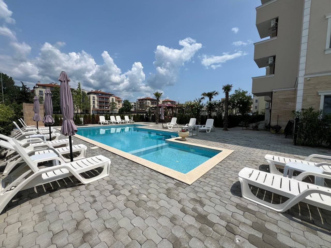 Современный бассейн с шезлонгами в жилом комплексе Болгарии, окруженный зелеными насаждениями и жилыми зданиями.