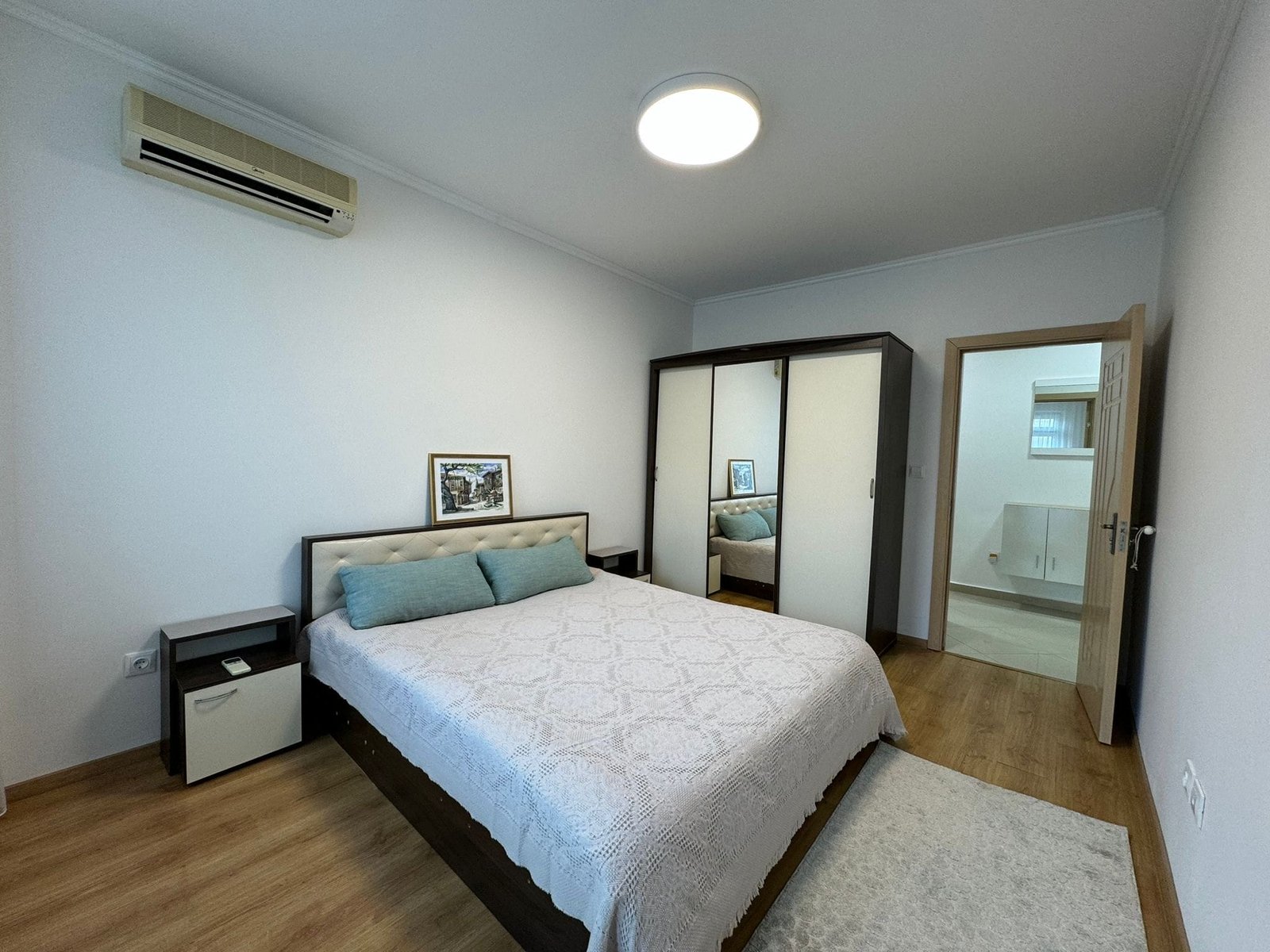 Современная спальня с двуспальной кроватью и зеркальным шкафом Современная спальня с двуспальной кроватью, шкафом, светлым интерьером и кондиционером, идеальная для покупки недвижимости в Болгарии.