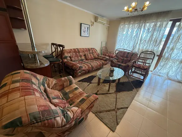 Современный интерьер гостиной с уютной мебелью, стеклянным столом и светлыми шторами, идеальный для покупки недвижимости в Болгарии.