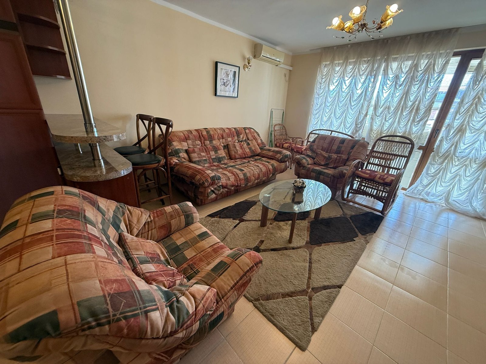 Современный интерьер гостиной с уютной мебелью, стеклянным столом и светлыми шторами, идеальный для покупки недвижимости в Болгарии.