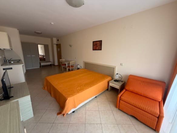 Студия в Болгарии с оранжевым покрывалом и диваном, светлые стены, кухня и зона для отдыха, идеальная для покупки недвижимости.