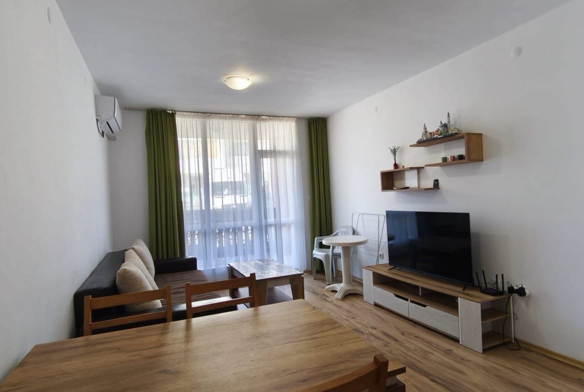 Современная гостиная с мягкой мебелью, телевизором, столом и балконом, оформленная в светлых тонах для комфортной жизни в Болгарии.