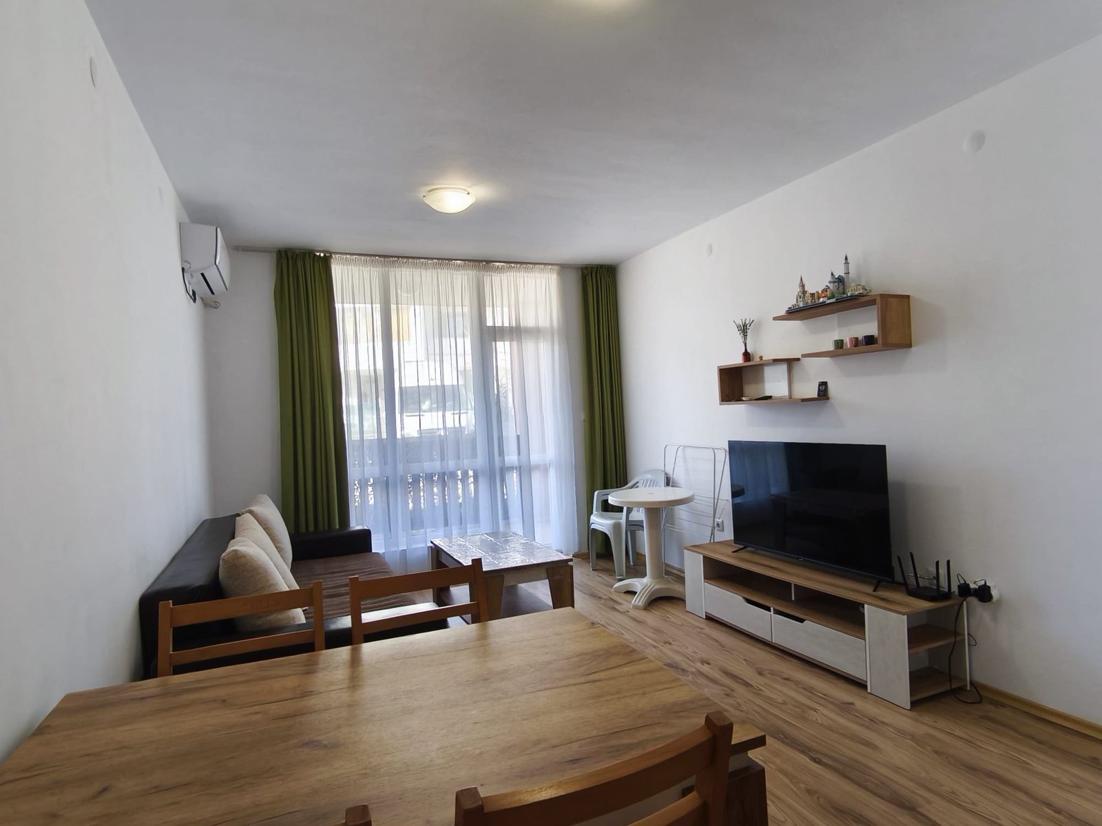 Современная гостиная с уютной мебелью и светлыми шторами Современная гостиная с мягкой мебелью, телевизором, столом и балконом, оформленная в светлых тонах для комфортной жизни в Болгарии.