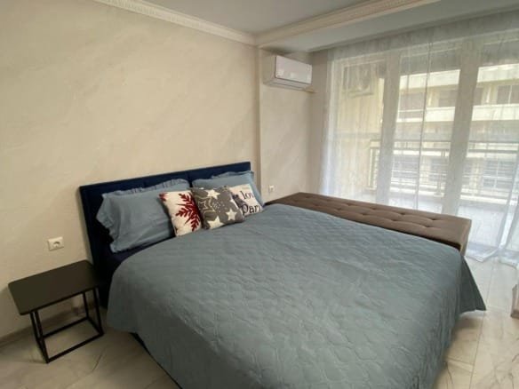 Современная спальня с двуспальной кроватью, светлыми тонами и уютной атмосферой, подходящая для продажи недвижимости в Болгарии.