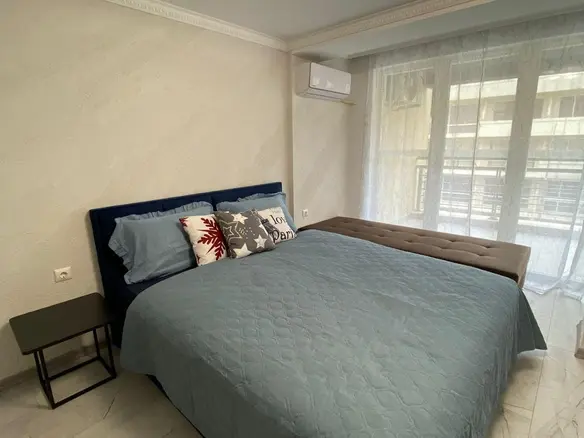 Современная спальня с двуспальной кроватью, светлыми тонами и уютной атмосферой, подходящая для продажи недвижимости в Болгарии.