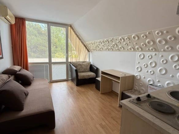 Современная студия с мягким диваном, креслом и кухонным уголком, большими окнами, светлое дерево на полу.