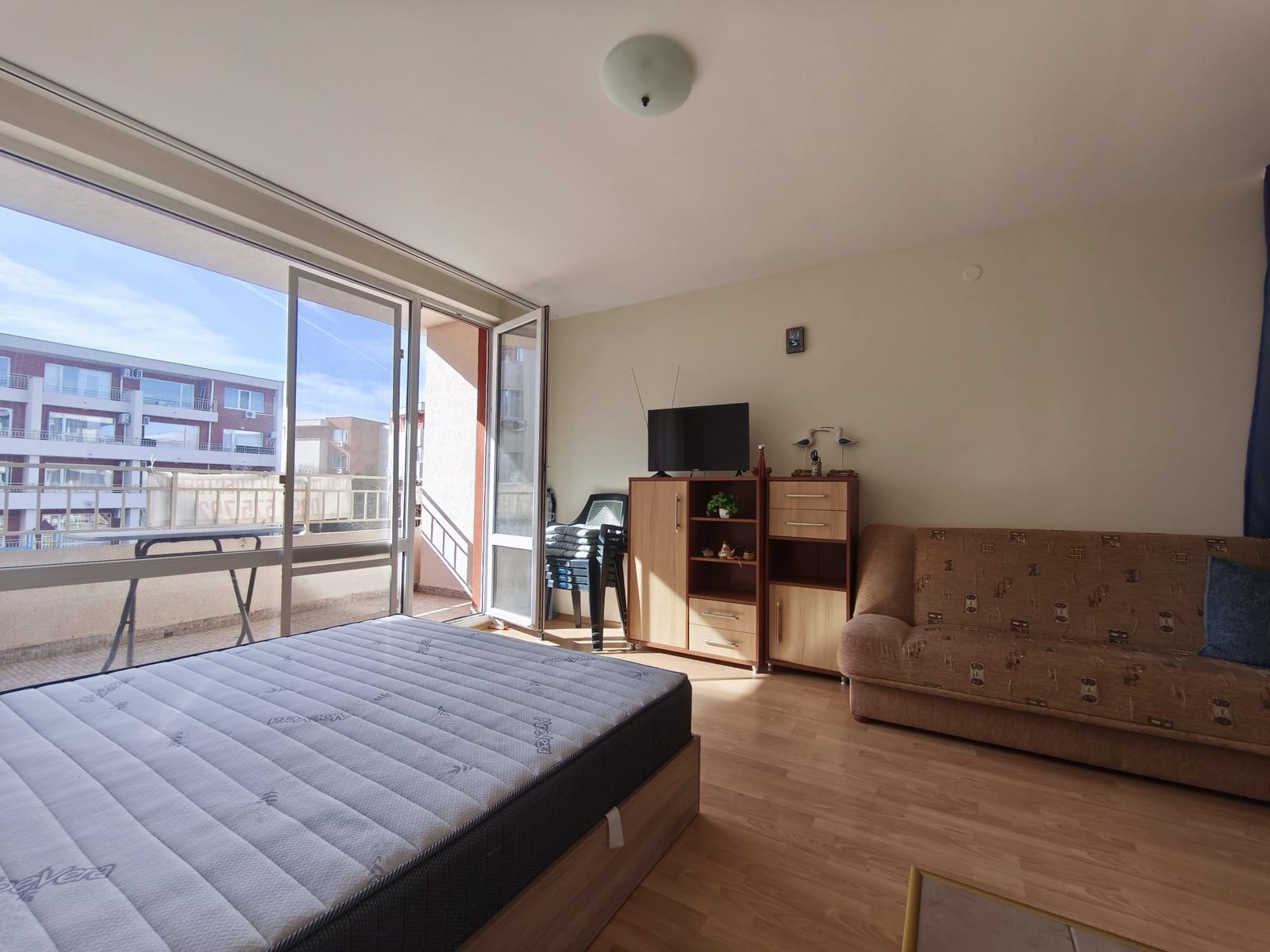 Уютная квартира с балконом и телевизором на стене Современная квартира с балконом в Болгарии, светлая спальня и уютная мебель для комфортного проживания.