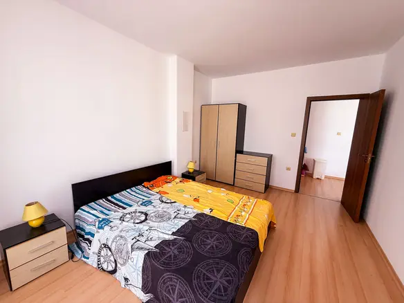 Современная спальня с большим двуспальным кроватью, комодом и шкафом, оформленная в светлых тонах для продажи недвижимости в Болгарии.