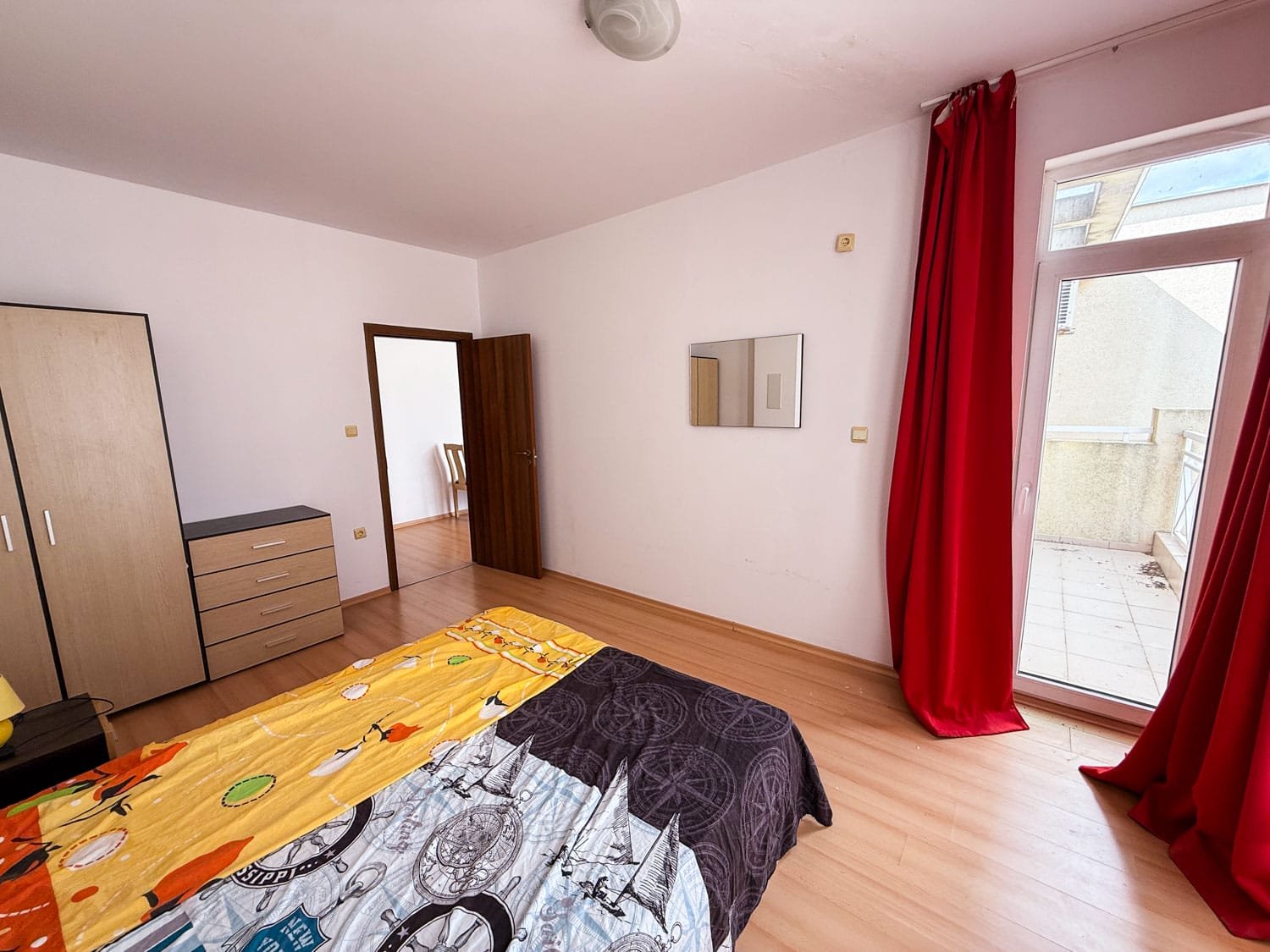 Светлая спальня с балконом, двуспальной кроватью с ярким текстилем и шкафом, предлагающая комфортное жилье в Болгарии.