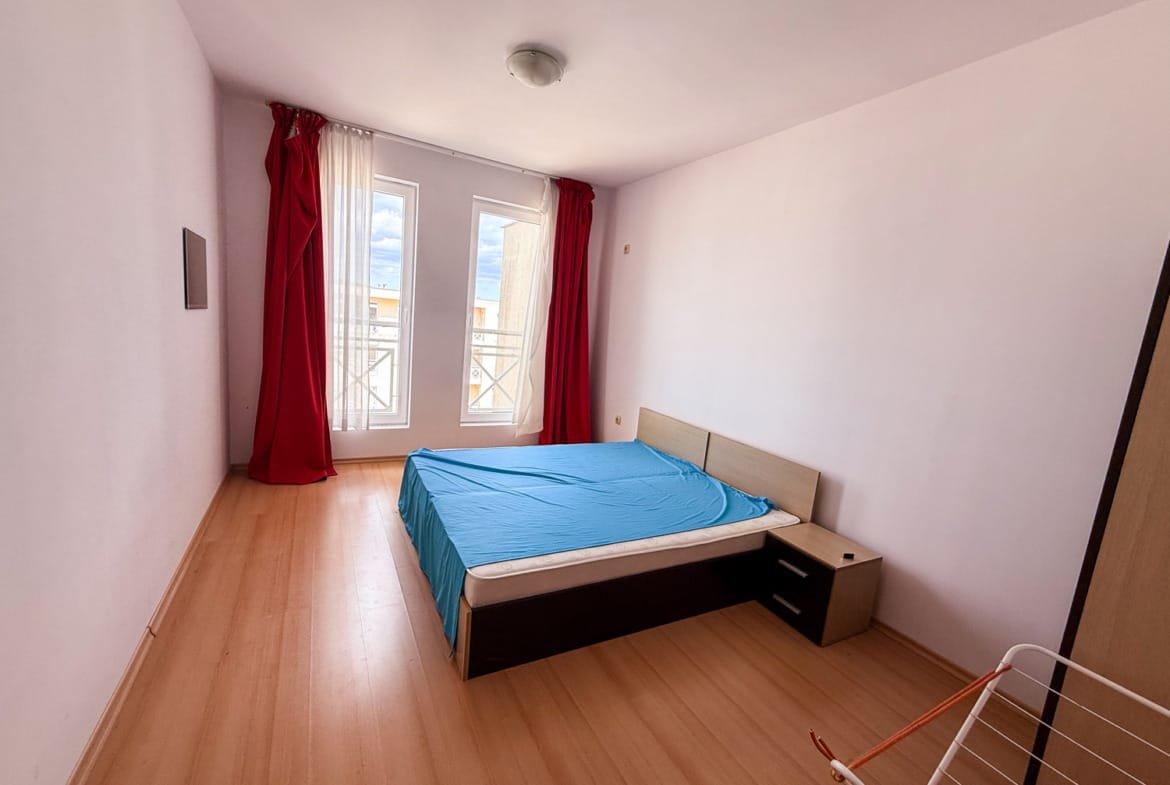 Светлая спальня с большие окнами, кроватью с синим покрывалом и красными занавесками, идеальна для покупки недвижимости в Болгарии.