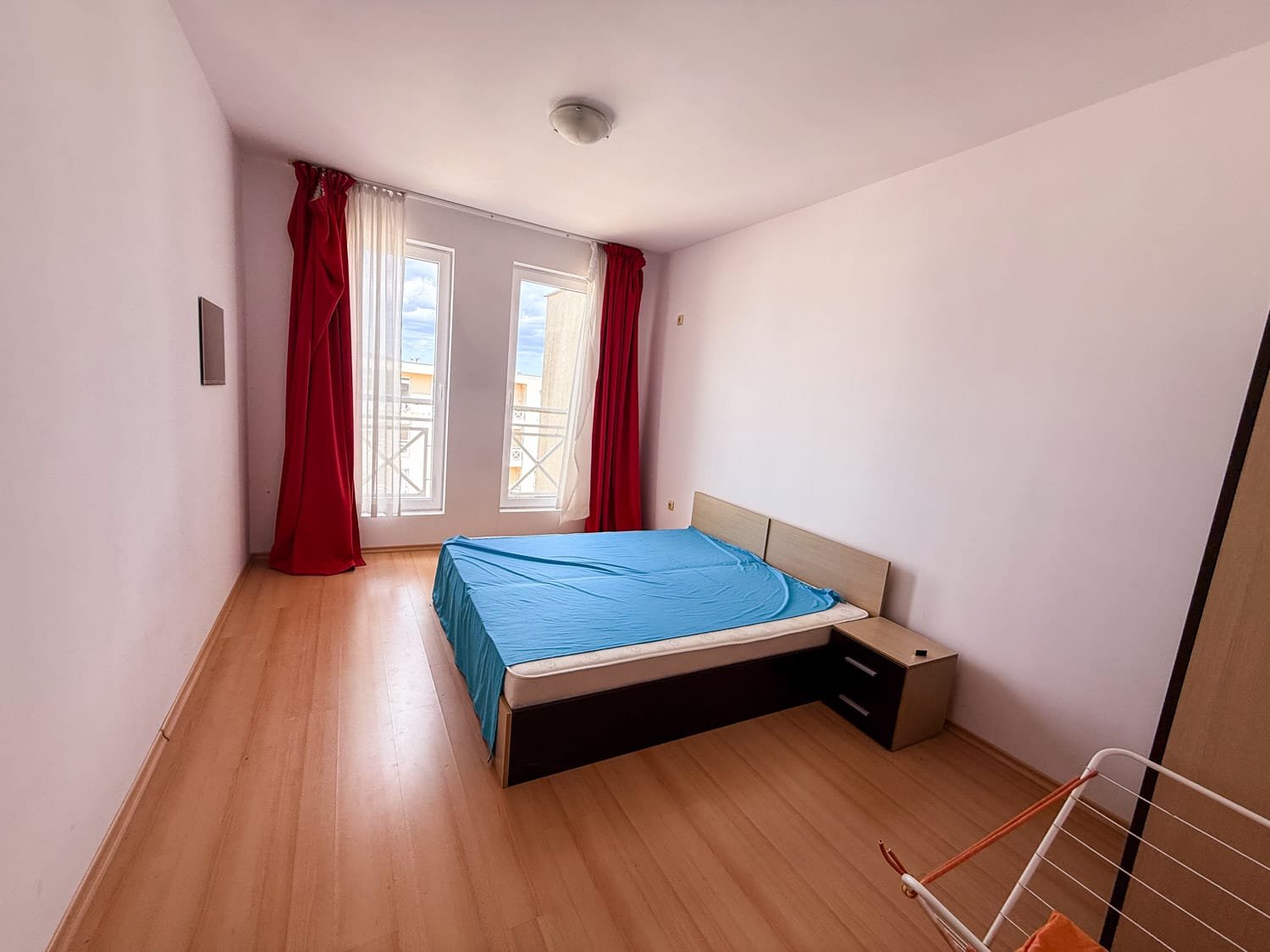 Светлая спальня с большие окнами, кроватью с синим покрывалом и красными занавесками, идеальна для покупки недвижимости в Болгарии.