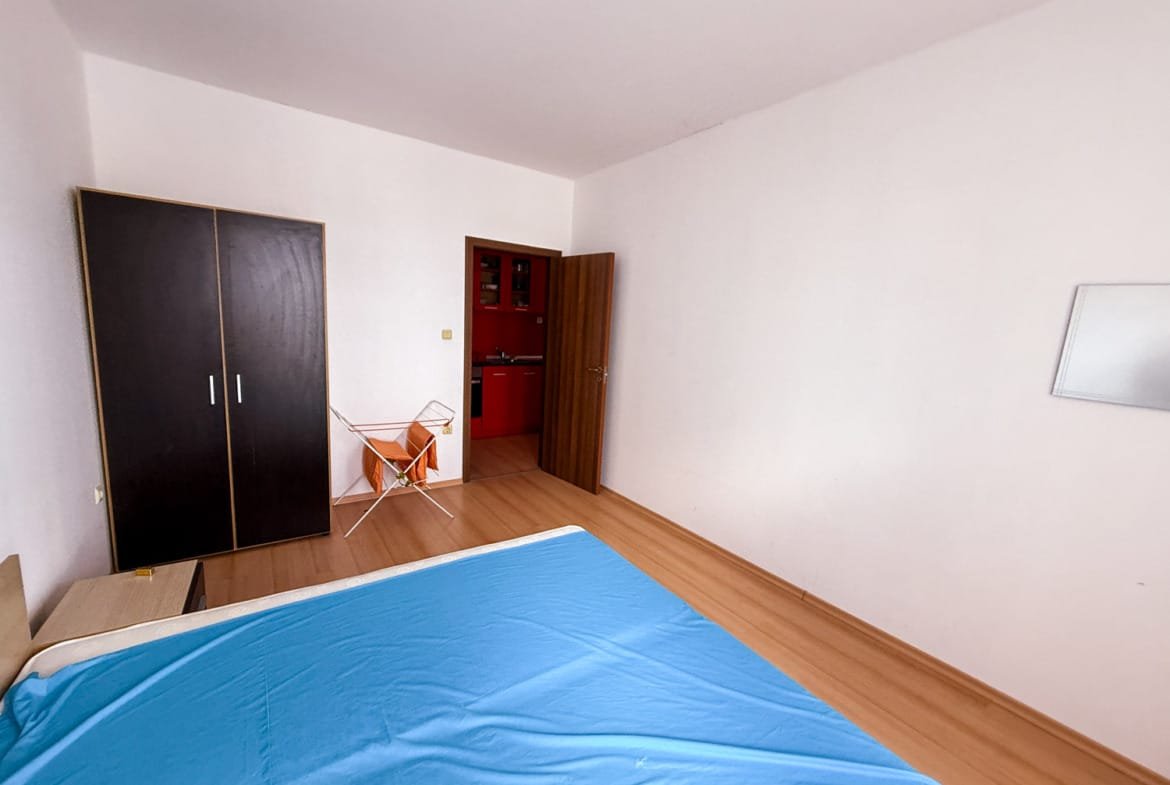 Современная спальня с односпальной кроватью, шкафом и кухней на фоне, идеально подходит для покупки недвижимости в Болгарии.