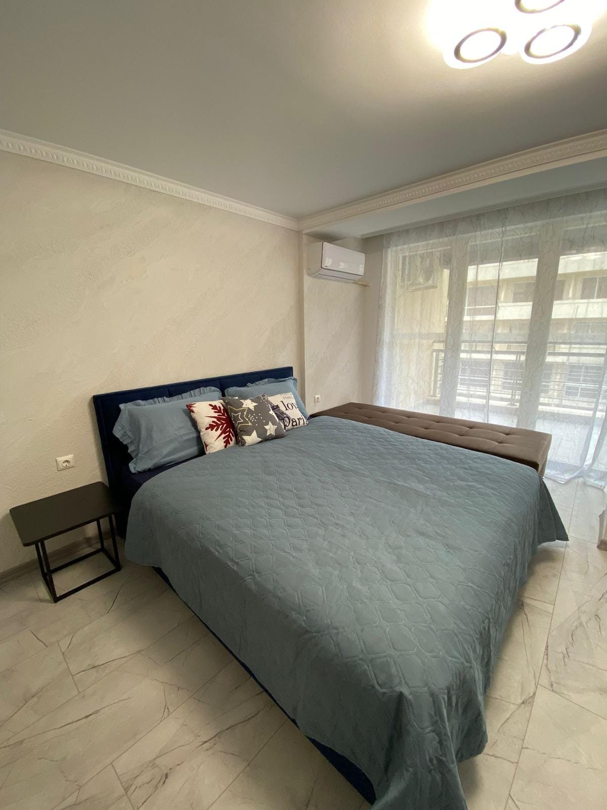 Современная спальня с двуспальной кроватью, светлыми тонами и уютной атмосферой, подходящая для продажи недвижимости в Болгарии.