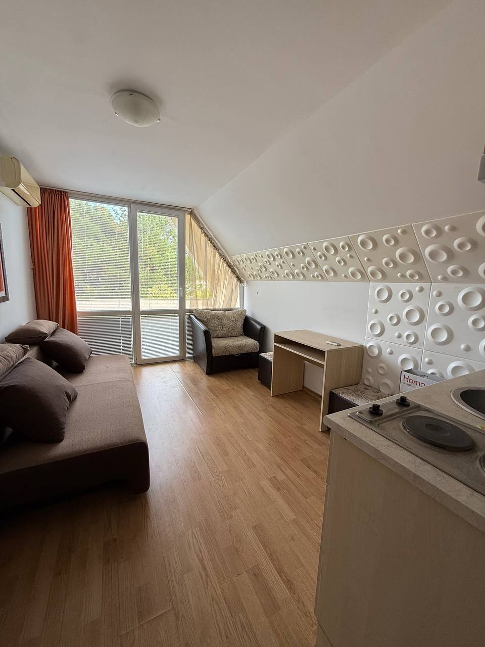 Современная студия с мягким диваном, креслом и кухонным уголком, большими окнами, светлое дерево на полу.