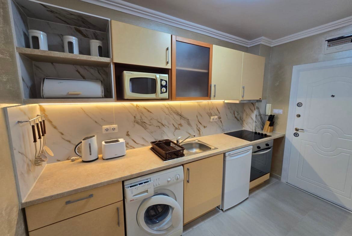 Современная кухня с встроенной техникой, стиральной машиной и плитой, оформленная в светлых тонах, для продажи недвижимости в Болгарии.