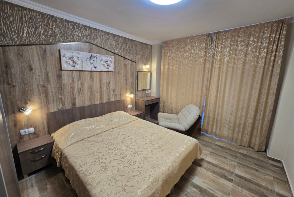 Современная спальня с двуспальной кроватью, деревянными элементами и уютными шторами в золотистых тонах. Подходит для аренды в Болгарии.