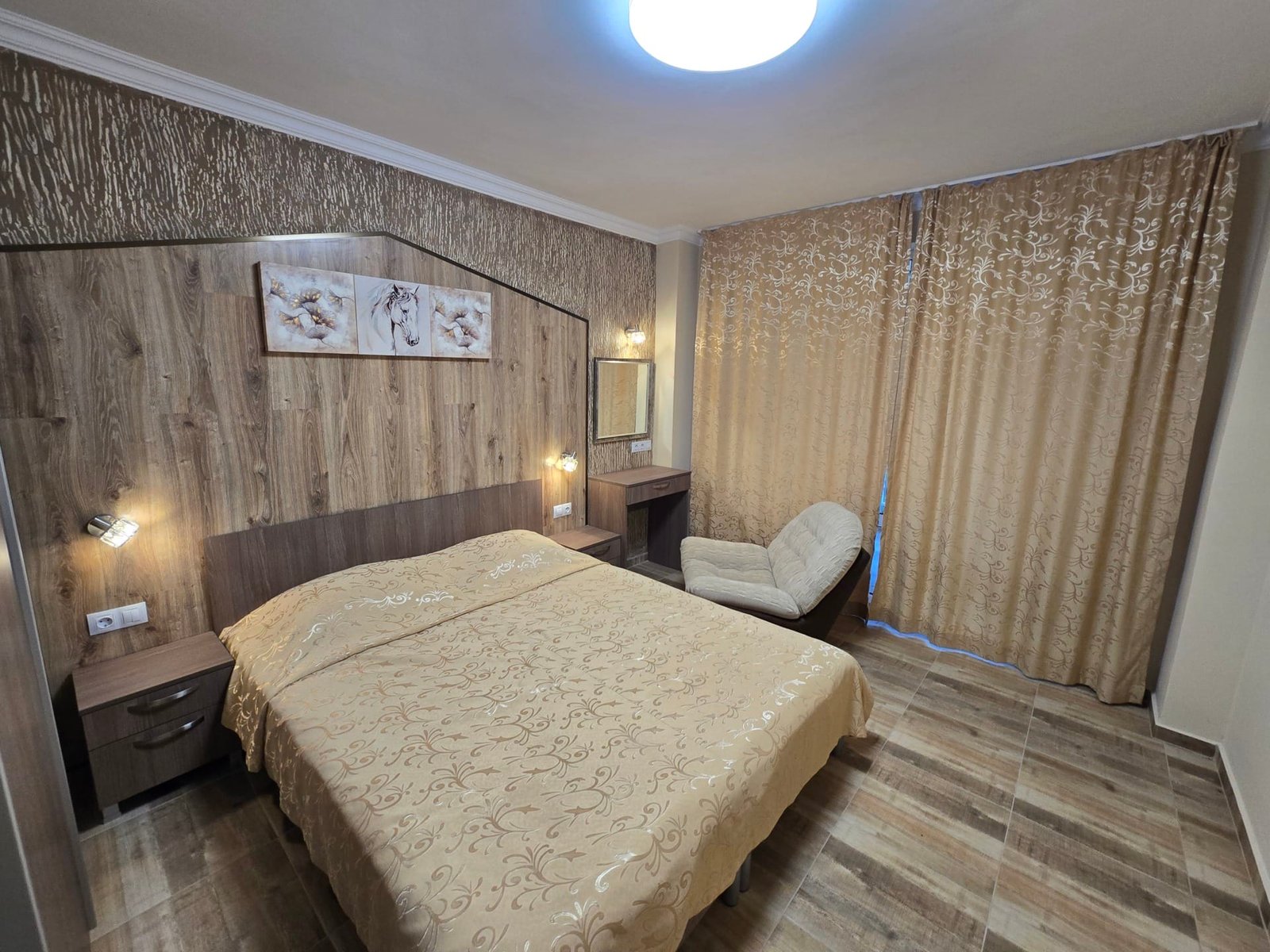 Современная спальня с двуспальной кроватью, деревянными элементами и уютными шторами в золотистых тонах. Подходит для аренды в Болгарии.