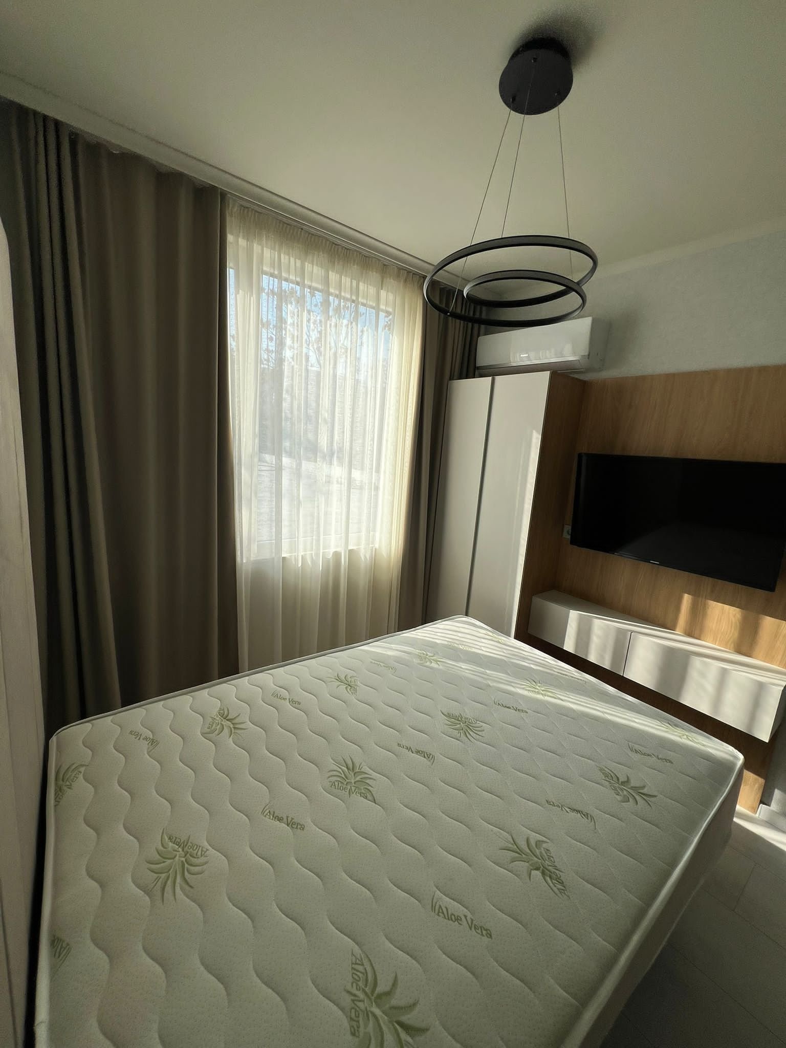 Современная спальня с белым матрасом, светлыми шторами и деревянной мебелью, идеальная для покупки недвижимости в Болгарии.