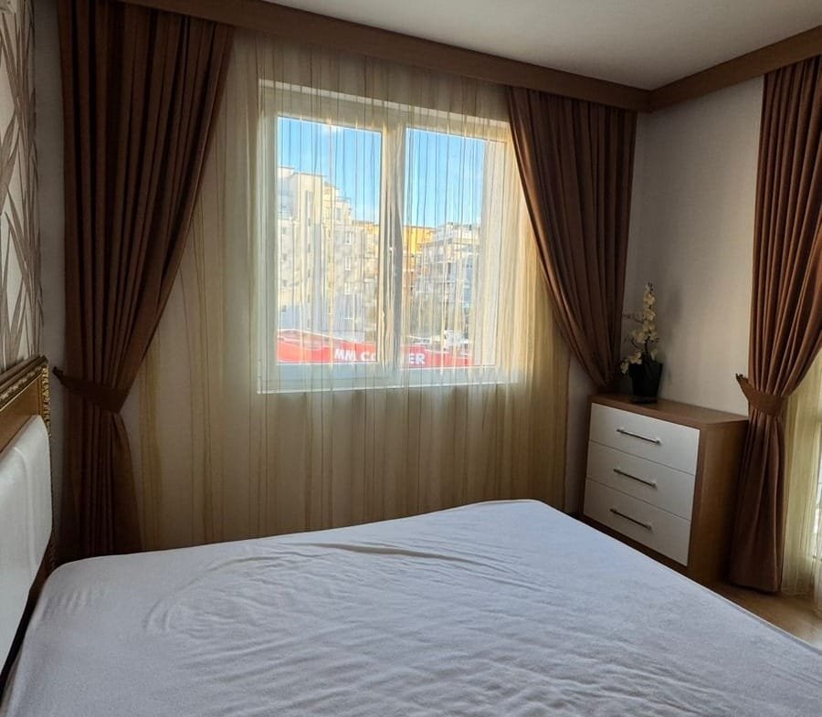 Светлая спальня с большими окнами, светлыми шторами и современной мебелью, идеальная для продажи недвижимости в Болгарии.