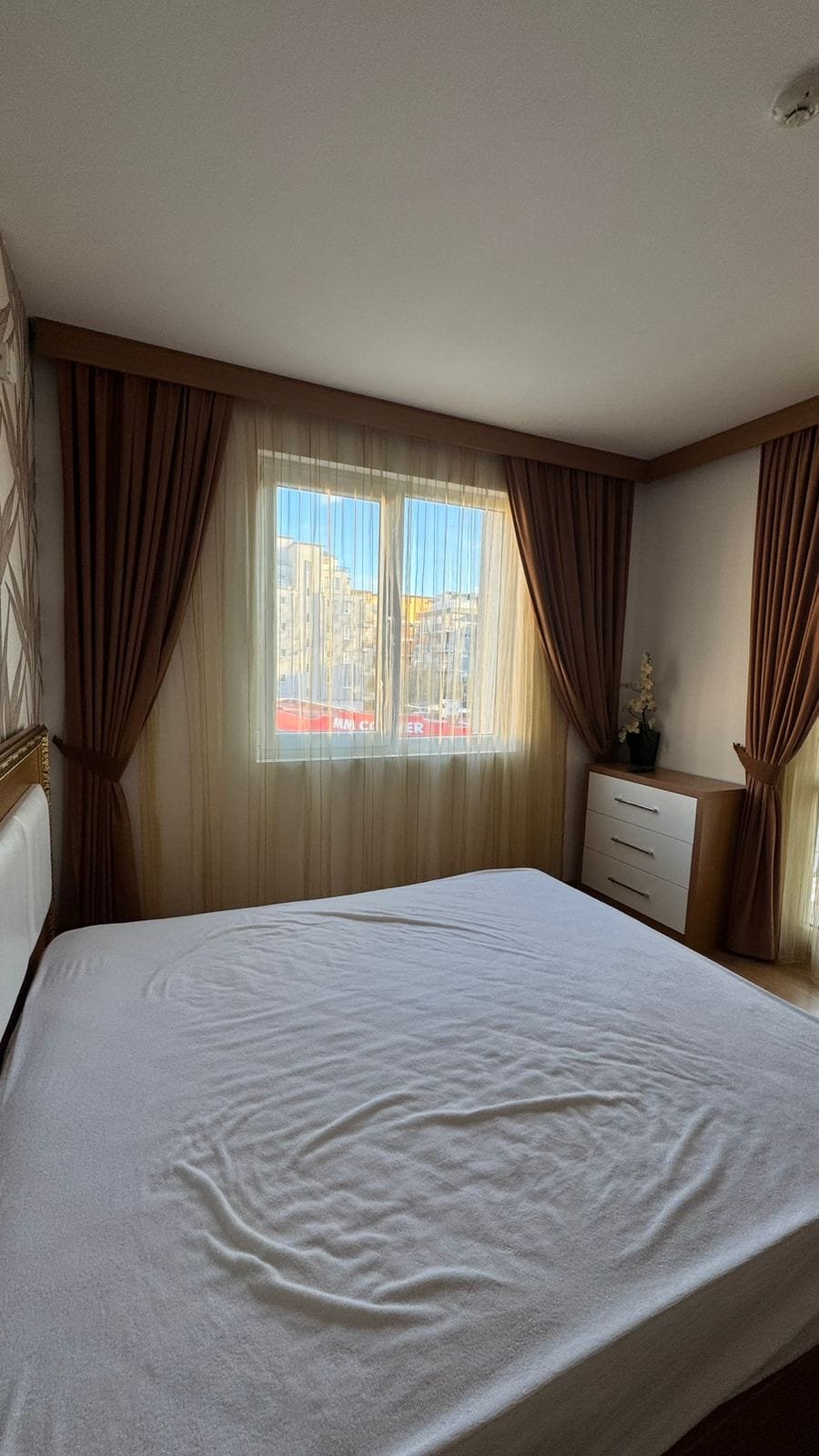 Светлая спальня с большими окнами, светлыми шторами и современной мебелью, идеальная для продажи недвижимости в Болгарии.