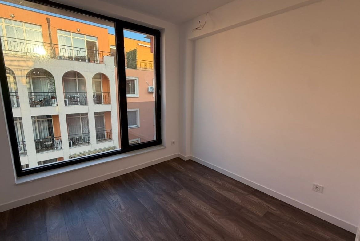 Современная комната с большим окном и видом на жилой комплекс в Болгарии, идеально подходящая для покупки недвижимости.