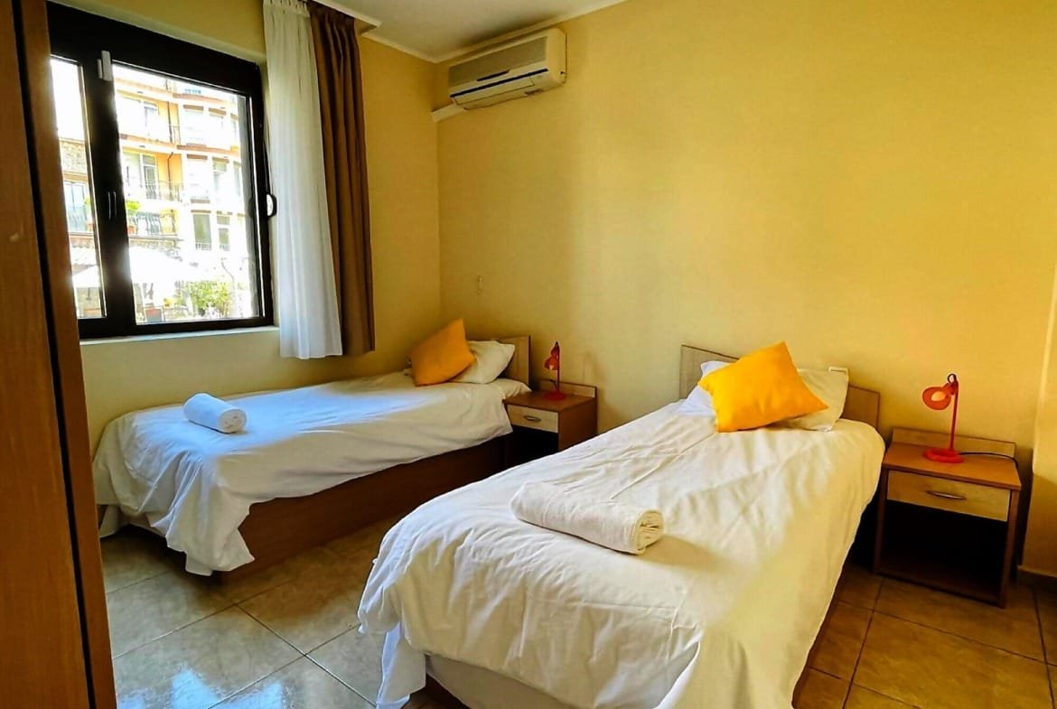 Современная спальня с двумя односпальными кроватями, светлыми стенами и акцентными подушками, идеальная для отдыха в Болгарии.