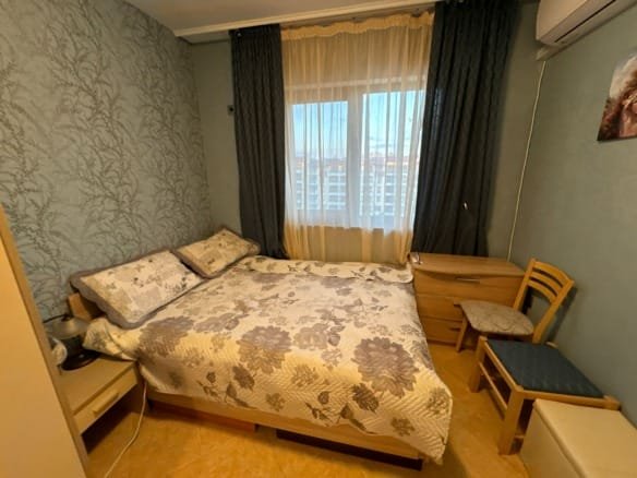 Комфортная спальня с двуспальной кроватью, тумбочкой и стулом, светлыми занавесками и приглушенными тонами, подходящая для жилья в Болгарии.