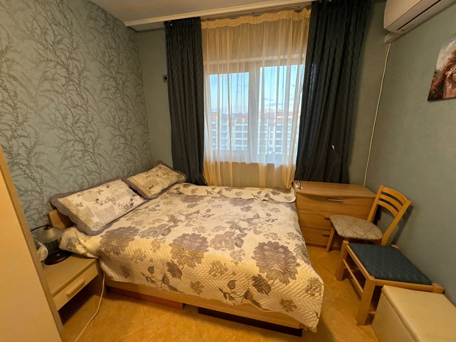 Уютная спальня с двуспальной кроватью и большим окном Комфортная спальня с двуспальной кроватью, тумбочкой и стулом, светлыми занавесками и приглушенными тонами, подходящая для жилья в Болгарии.