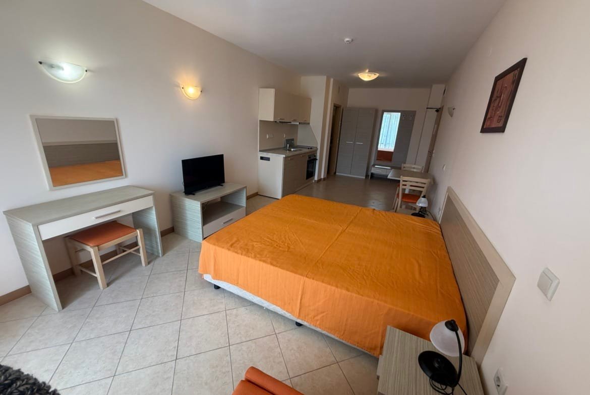 Современная квартира с оранжевым покрывалом, телевизором, кухонной зоной и рабочим местом. Идеально для покупки недвижимости в Болгарии.