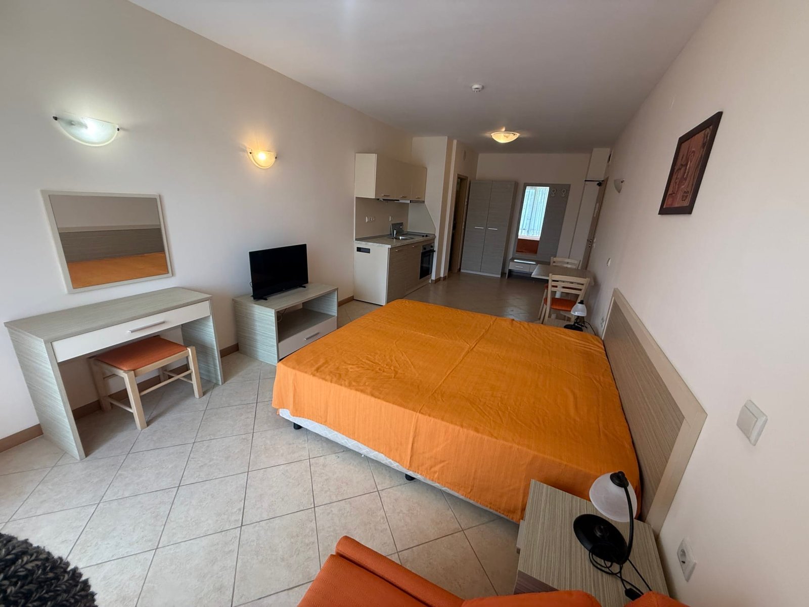 Современная квартира с оранжевым покрывалом, телевизором, кухонной зоной и рабочим местом. Идеально для покупки недвижимости в Болгарии.