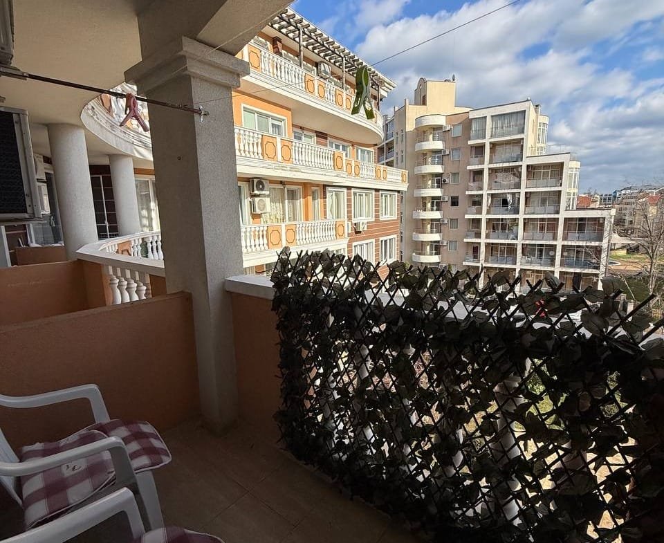 Балкон с видом на город, украшенный зелеными растениями и двумя белыми стульями на фоне жилых зданий и голубого неба.