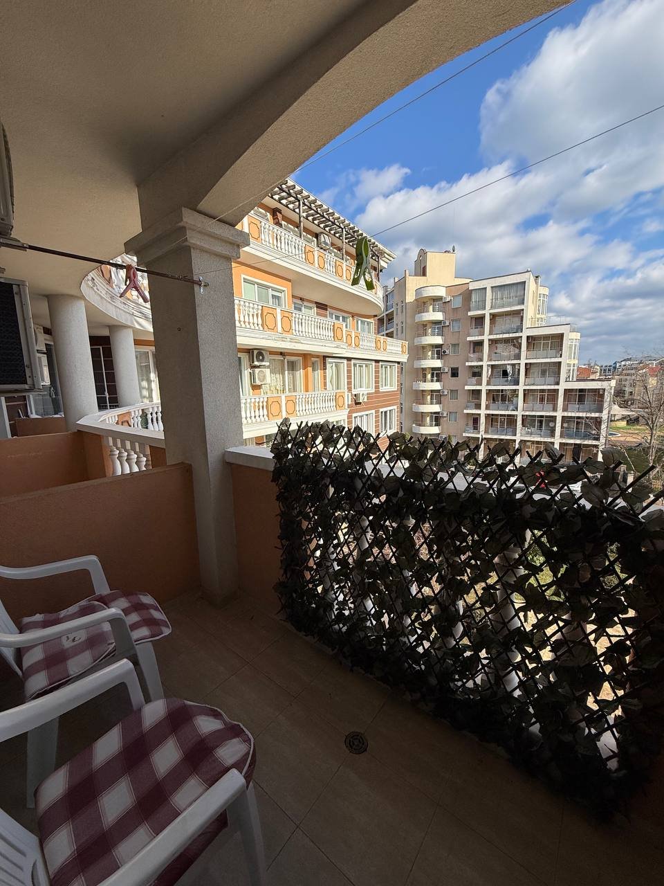 Балкон с видом на город, украшенный зелеными растениями и двумя белыми стульями на фоне жилых зданий и голубого неба.