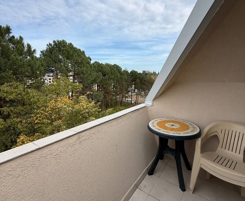 Уютный балкон с круглым столом и пластиковым стулом, видом на зелёные деревья и спокойную атмосферу загородного комплекса.