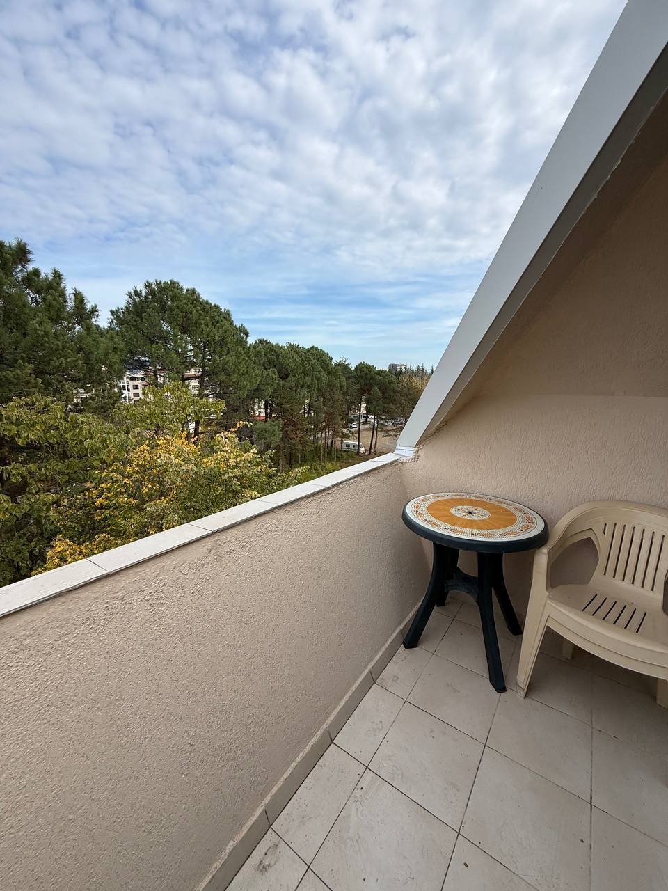 Уютный балкон с круглым столом и пластиковым стулом, видом на зелёные деревья и спокойную атмосферу загородного комплекса.