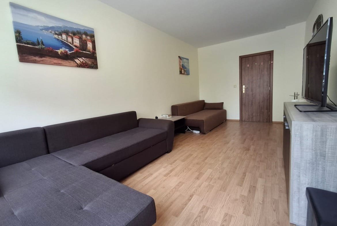 Современная гостиная с тёмно-коричневыми диванами, деревянным полом и телевизором на стене, украшенная картинами.