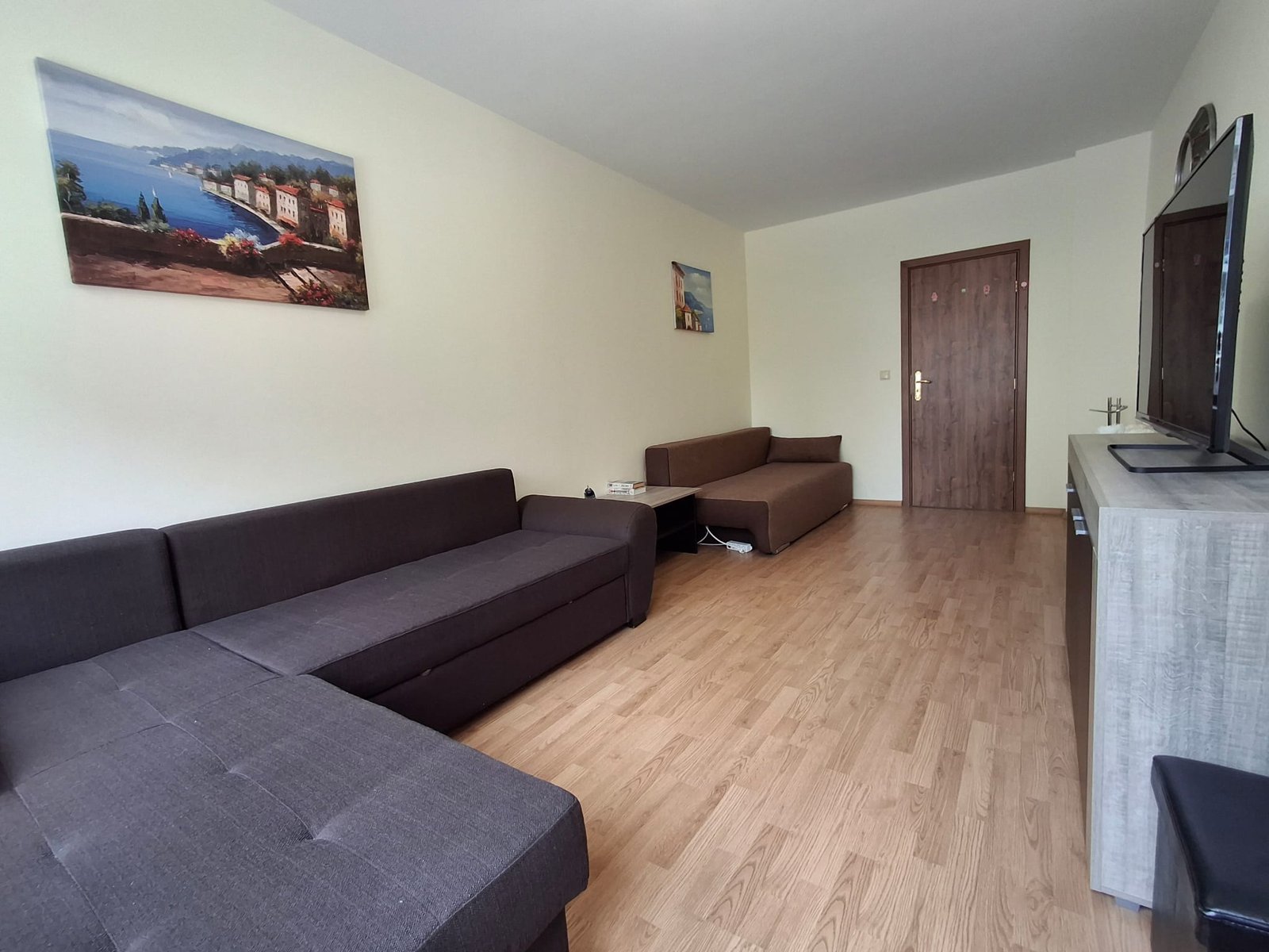 Современная гостиная с тёмно-коричневыми диванами, деревянным полом и телевизором на стене, украшенная картинами.