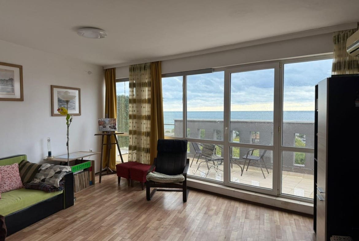 Современная гостиная с панорамным окном, видом на море и выходом на террасу; уютная обстановка для отдыха в Болгарии.