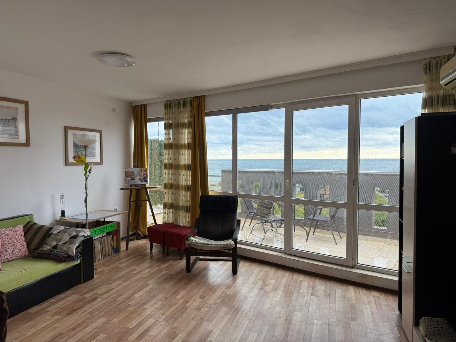 Современная гостиная с панорамным окном, видом на море и выходом на террасу; уютная обстановка для отдыха в Болгарии.