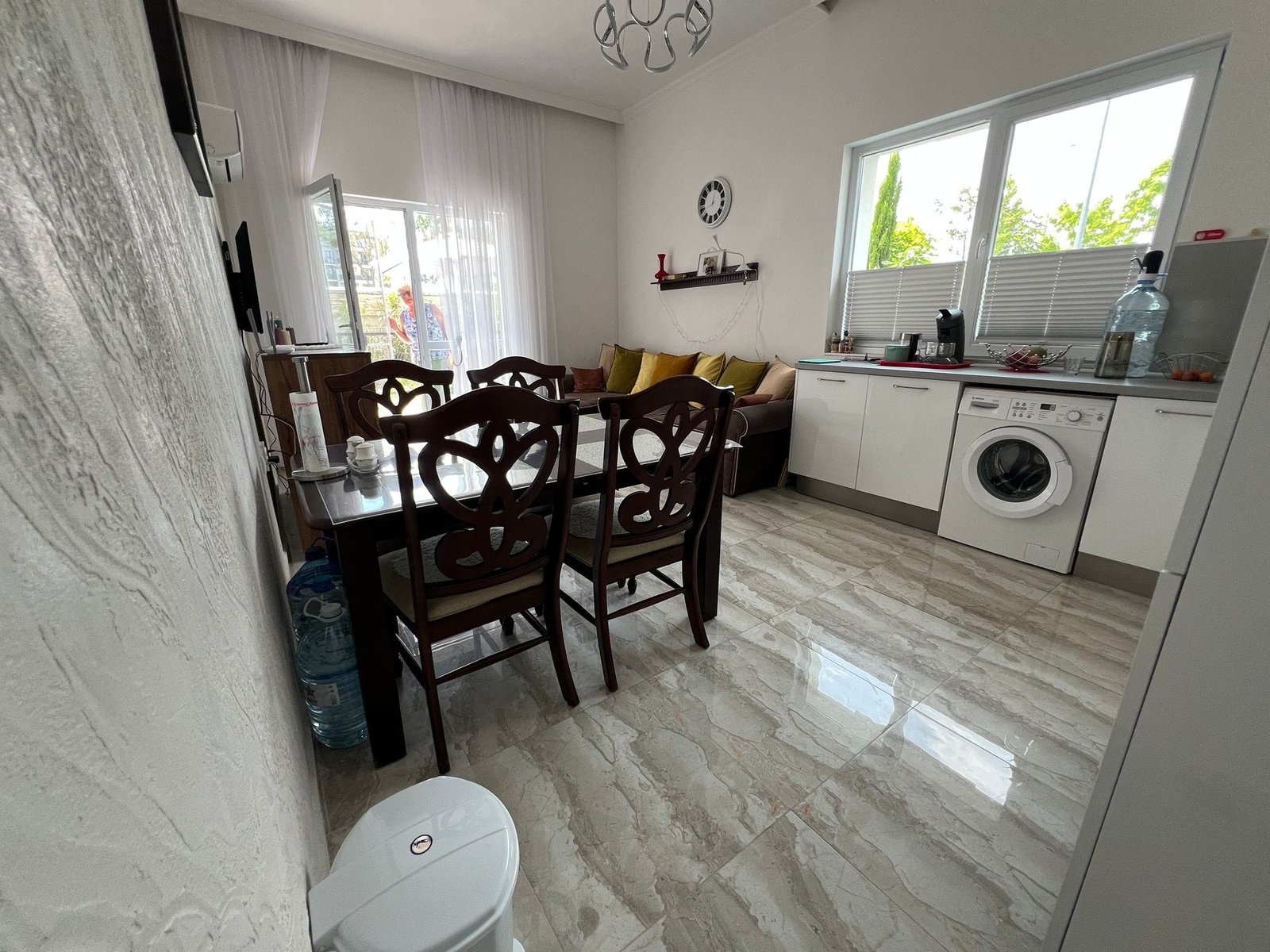 Уютная кухня-гостиная с обеденным столом, стиральной машиной и светлыми шторами, идеальная для покупки недвижимости в Болгарии.