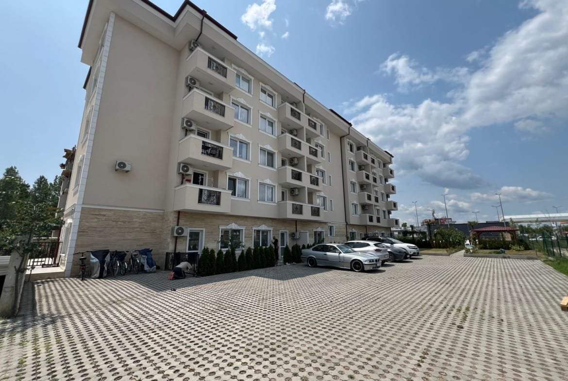 Современный многоквартирный дом с парковкой, расположенный в Болгарии, окруженный зелеными насаждениями и ясным небом.