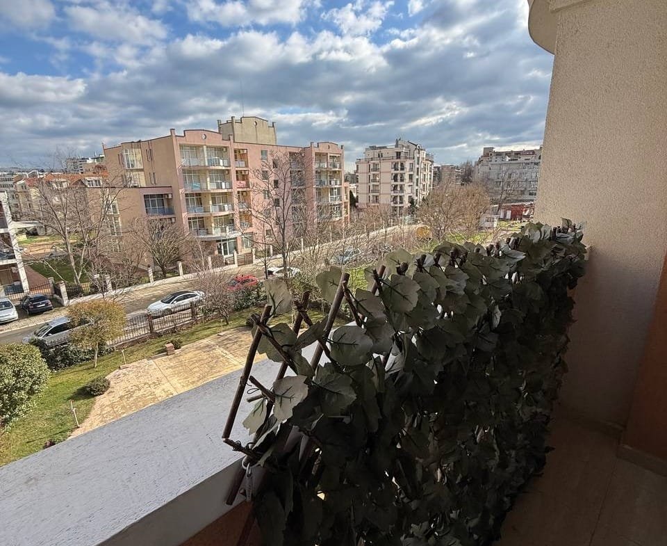 Балкон с зелеными растениями, вид на городские здания и ясное голубое небо с облаками.