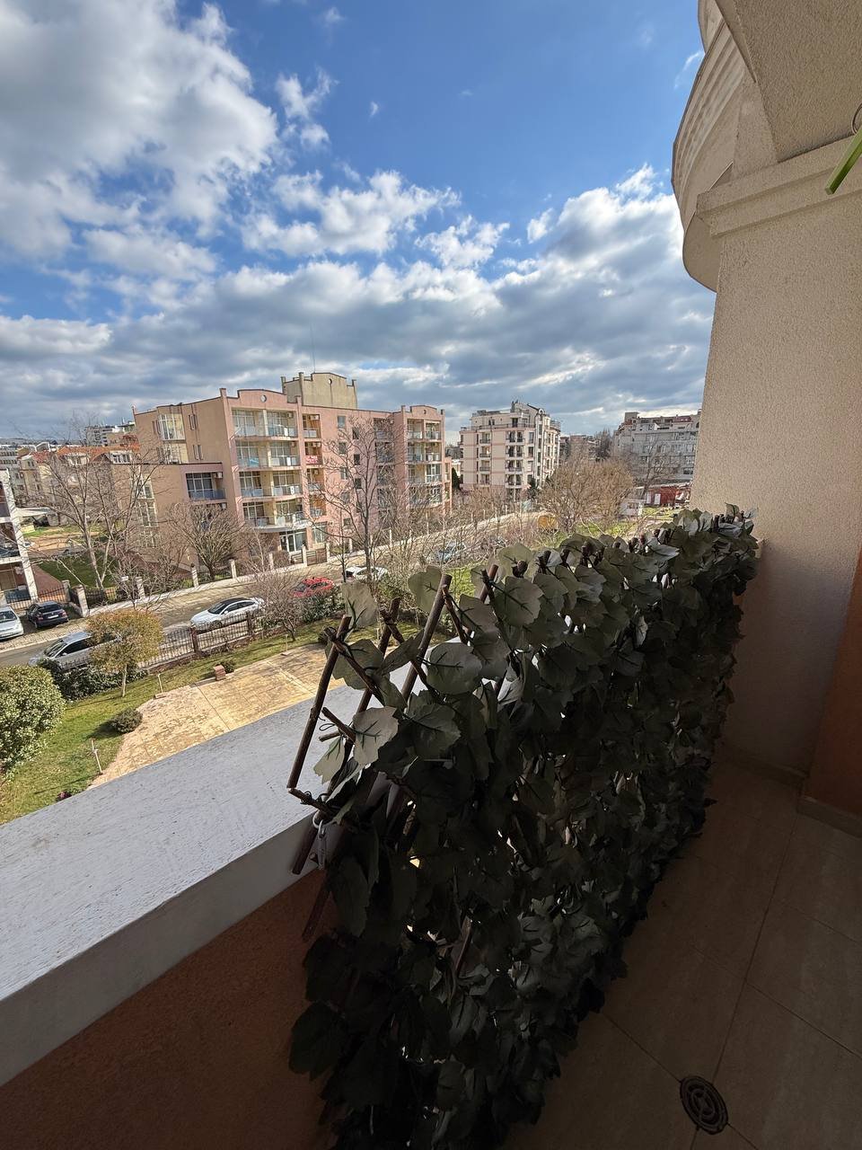 Балкон с зелеными растениями, вид на городские здания и ясное голубое небо с облаками.