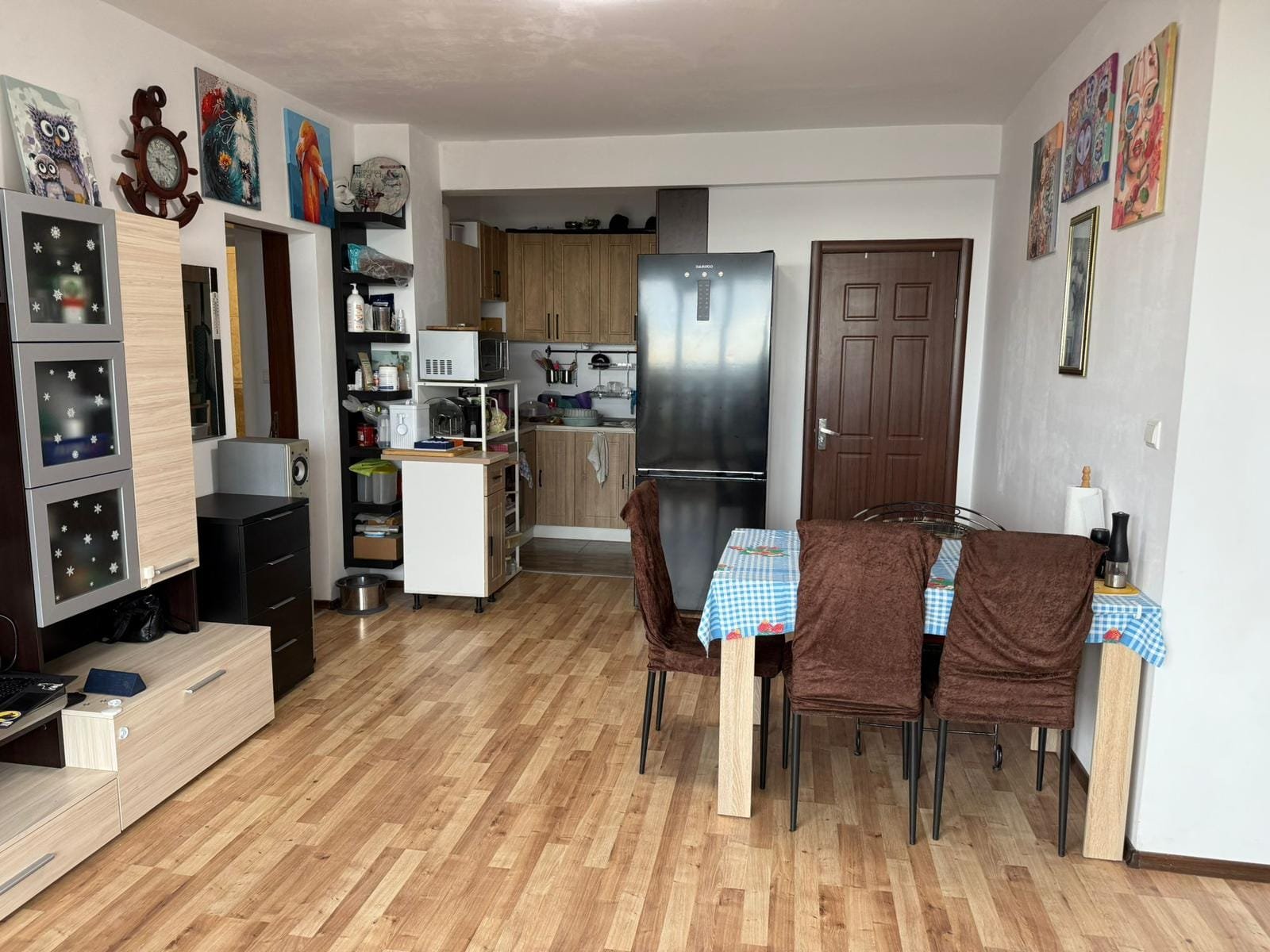 Современная кухня-гостиная с деревянным полом, обеденным столом, холодильником и яркими картинами на стенах в интерьере Болгарии.