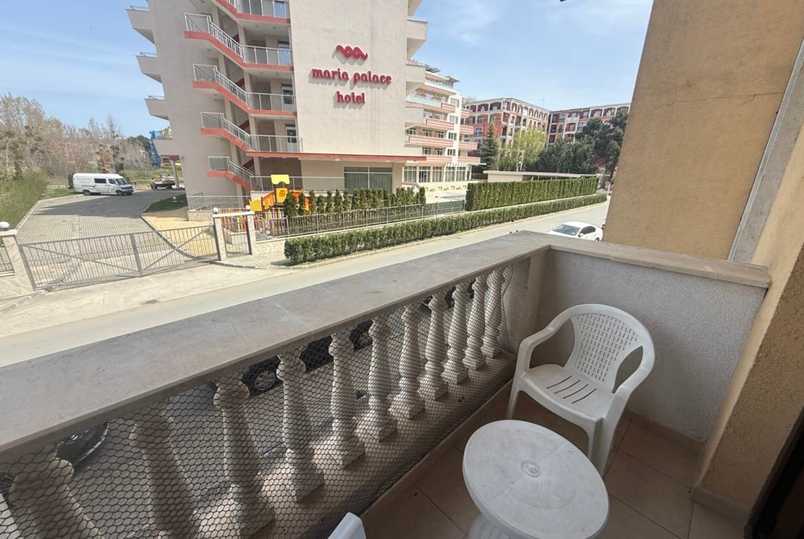 Балкон с пластиковыми стульями и столиком, вид на отель Maria Palace, зелёные насаждения и улица в Болгарии.