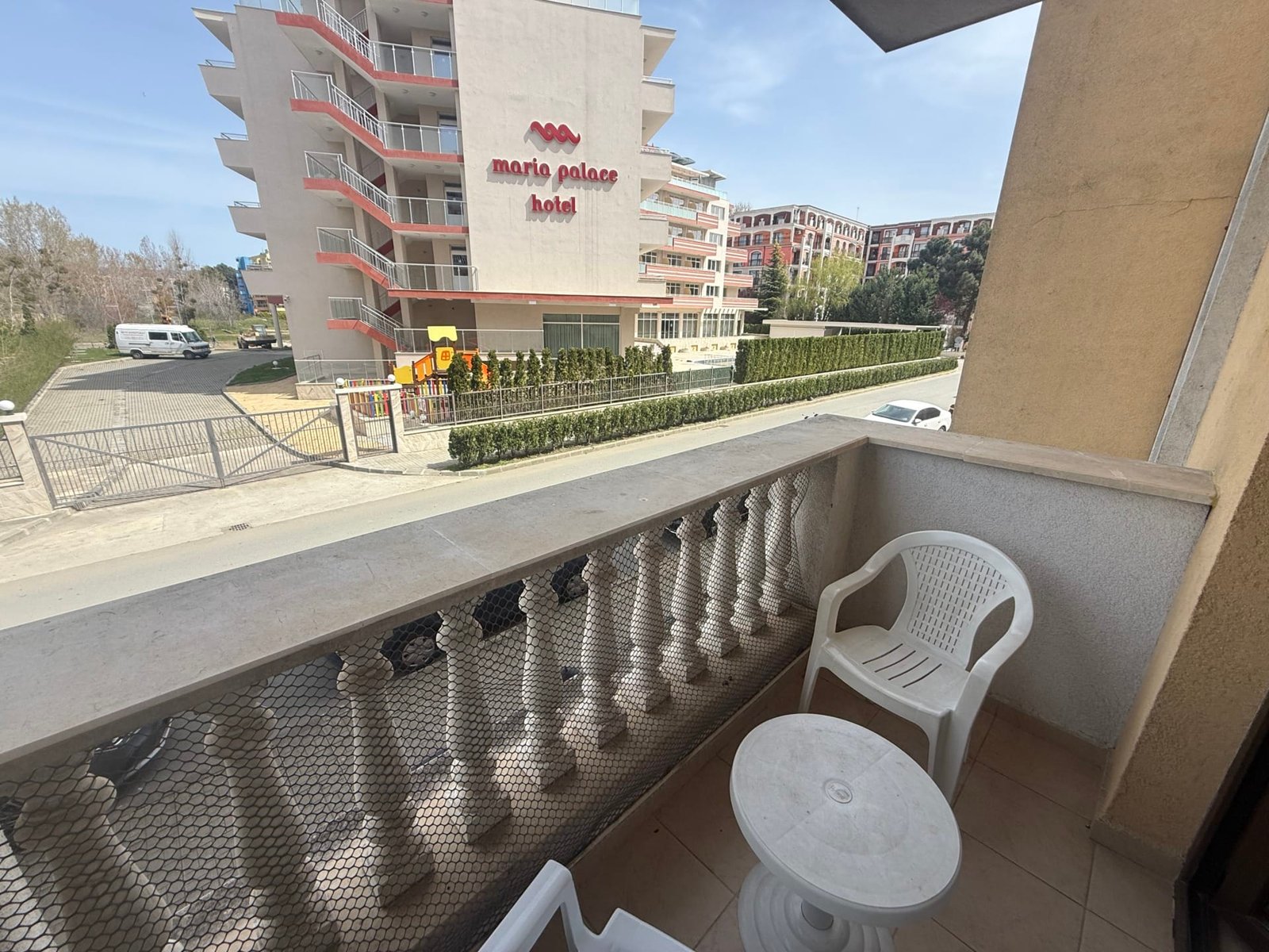 Балкон с пластиковыми стульями и столиком, вид на отель Maria Palace, зелёные насаждения и улица в Болгарии.